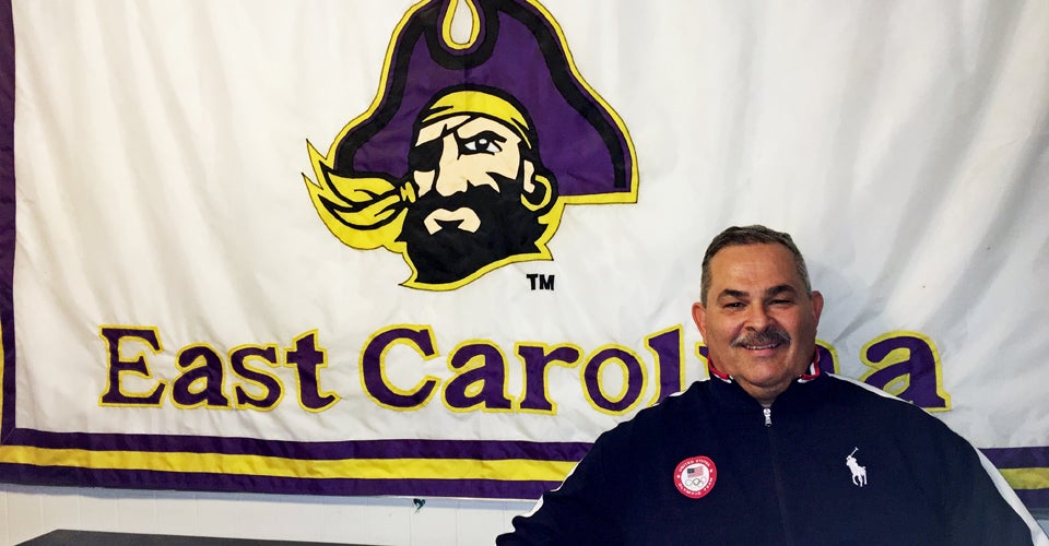 Tim Swords ’82 sports his Team USA jacket in front of an East Carolina banner at his weightlifting facility in Texas.