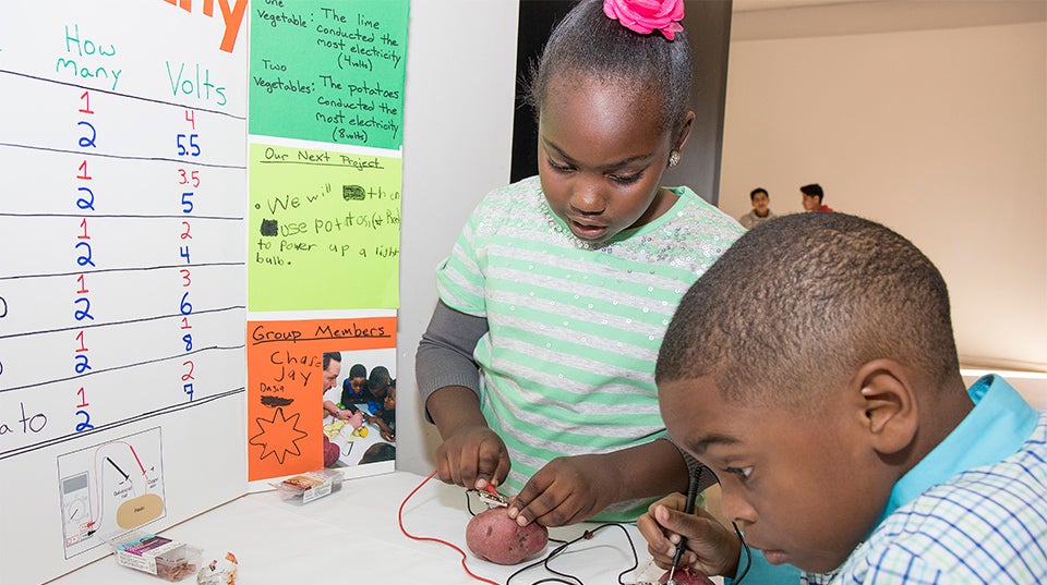 Clifton Daniels, age 6, and Dasia Little, age 8, demonstrate their experiment that measures the electricity produced by different vegetables. Clifton reads a voltage meter while both students connect probes to the potatoes.