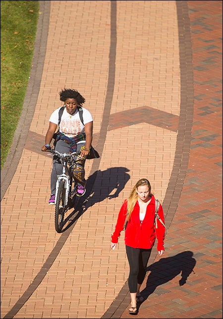 Using the marked bicycle lanes on campus, a student riding a bicycle travels alongside another student on foot. The bicycle lanes marked by different colored bricks were designed to enhance traffic on campus by allowing specific places for pedestrians and bicyclists.