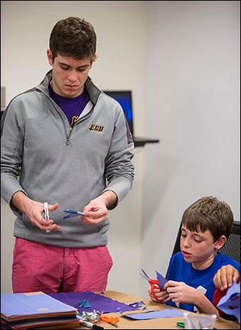 ECU student Patrick Young, standing, shows Nick Dixon how to make cuts to create a paper snowflake.