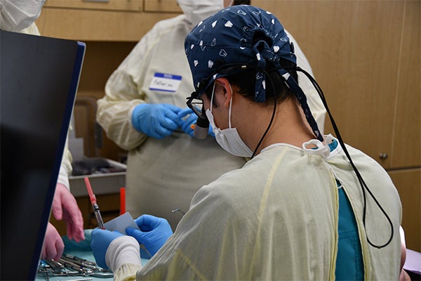 Dental care providers in protective equipment handle instruments in an operatory bay as they work on a patient’s teeth.