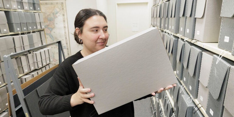 A woman with dark hair, wearing a black sweater, stands between shelving units lifting a box to add it to the shelf.