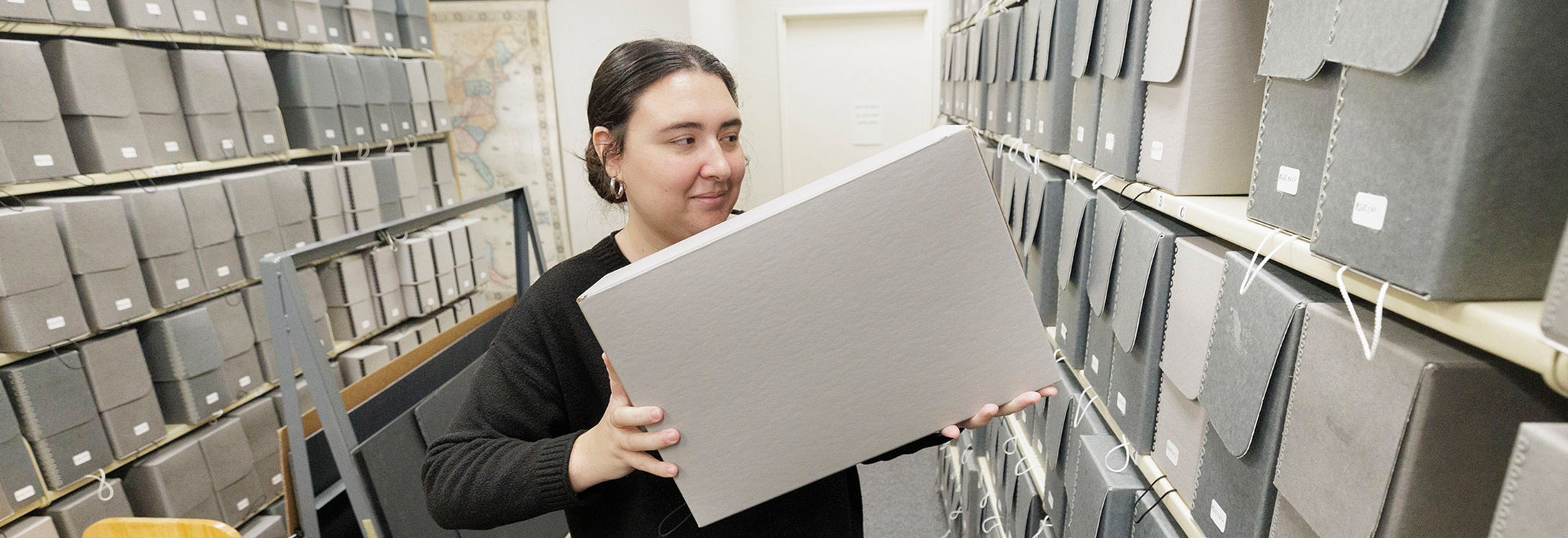 A woman with dark hair, wearing a black sweater, stands between shelving units lifting a box to add it to the shelf.