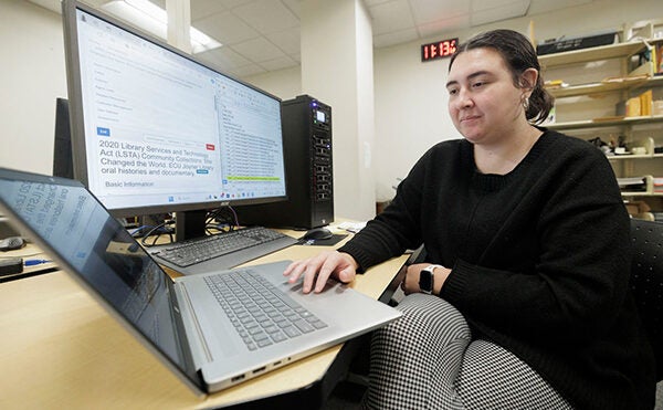 A woman sits at a table in front of a laptop computer and monitor.