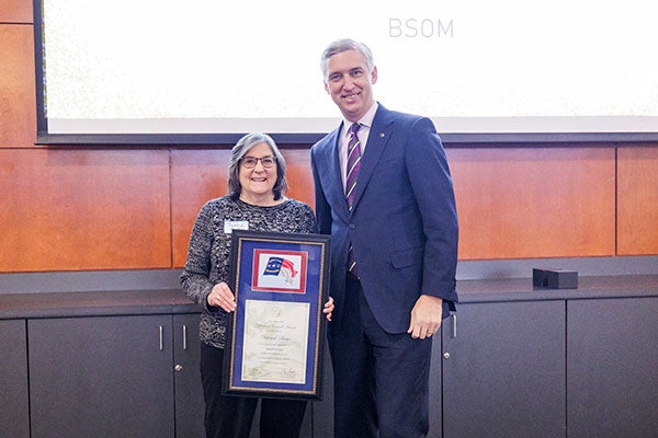 A woman with salt and pepper hair and wearing glasses is presented a framed certificate emblazoned with a state of North Carolina flag from a tall man in a blue suit and purple and gold tie. 