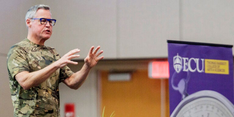 A man talks and gestures with his hands as he stands on stage in the Main Campus Student Center. There is a podium behind him and the Thomas Harriot College of Arts and Sciences banner beside him.