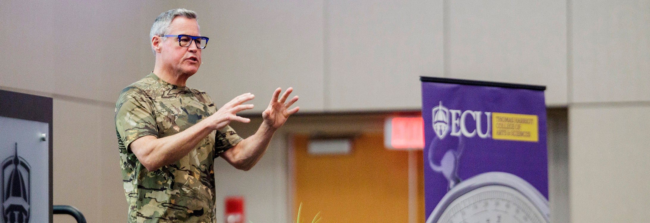 A man talks and gestures with his hands as he stands on stage in the Main Campus Student Center. There is a podium behind him and the Thomas Harriot College of Arts and Sciences banner beside him.