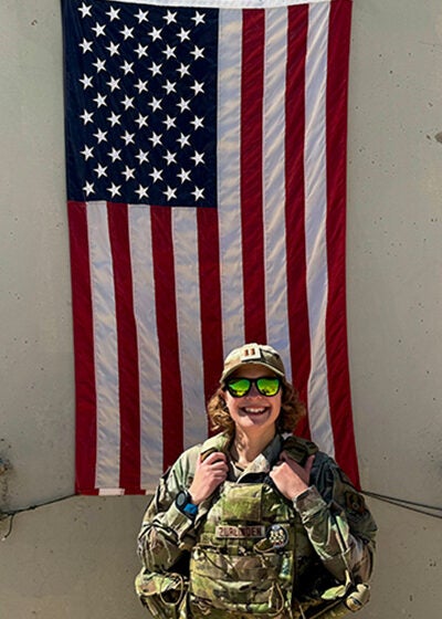 A woman wearing fatigues and sunglasses holds a tactical plate carrier across her chest. She is standing in front of a United States flag attached to the concrete wall behind her.