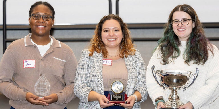 Three women stand in front of a white screen in East Carolina University’s Main Campus Student Center. They each hold a different trophy representing their first, second and third place awards in the Three-Minute Thesis competition.