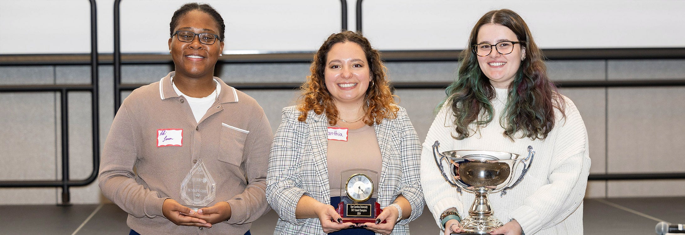 Three women stand in front of a white screen in East Carolina University’s Main Campus Student Center. They each hold a different trophy representing their first, second and third place awards in the Three-Minute Thesis competition.