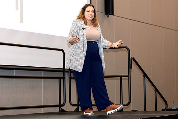 A woman in dark slacks, tan shirt and a gray plaid blazer talks on a stage in the Main Campus Student Center.