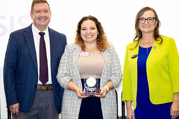 A woman in dark slacks, tan shirt and a gray plaid blazer holds a first-place trophy and stands in front of a table with a tablecloth advertising the Three-Minute Thesis competition.