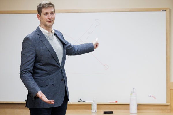 A man in a blue sport jacket points at a white board behind him.