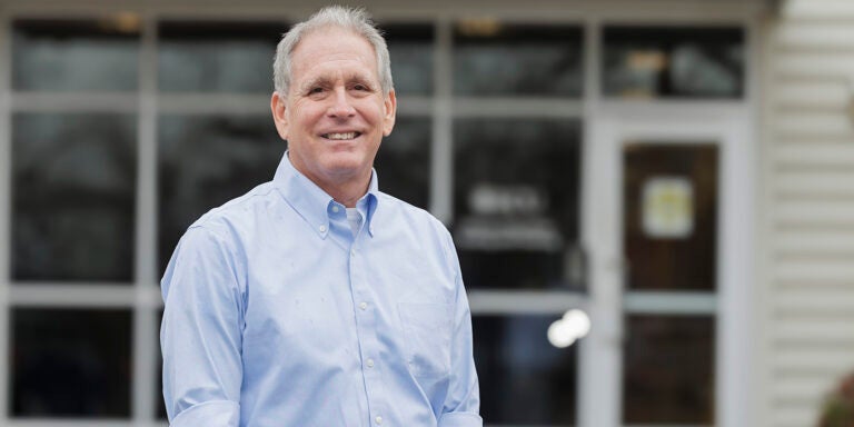 A man in blue shirt stands in front of a building, proudly smiling.