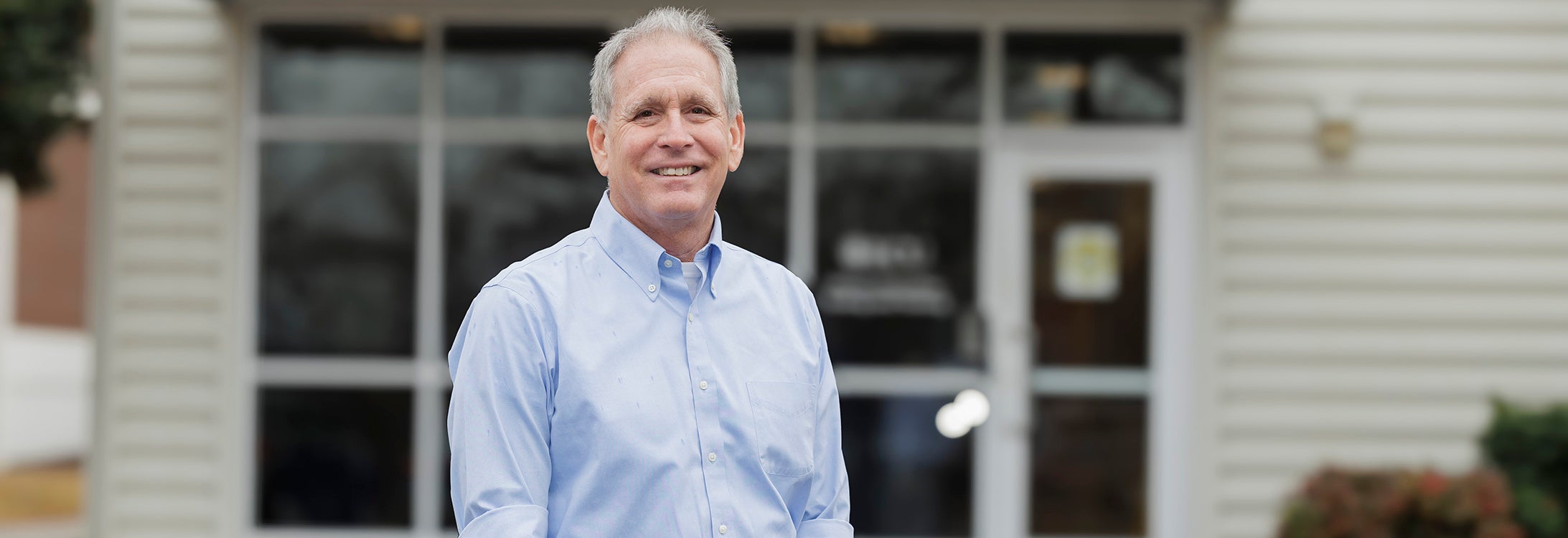 A man in blue shirt stands in front of a building, proudly smiling.