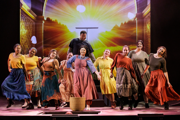 A group of ten performers in brightly colored wardrobes dance with their hands on their hips.