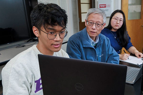 Three people sit at a table collaborating, with one student holding a laptop while the others look on and take notes in a classroom setting.