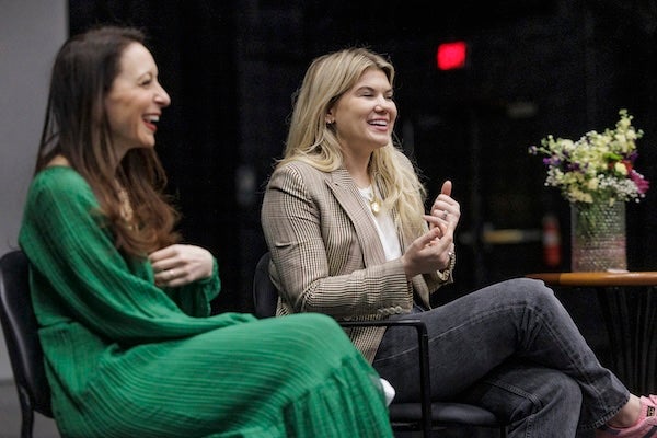 Two women sit in chairs next to each other on the floor of a theater. A large vase of flowers is on a background table.