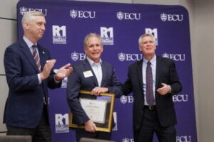 A man on the left applauds a smiling man in middle holding a framed award while a man on right gestures.