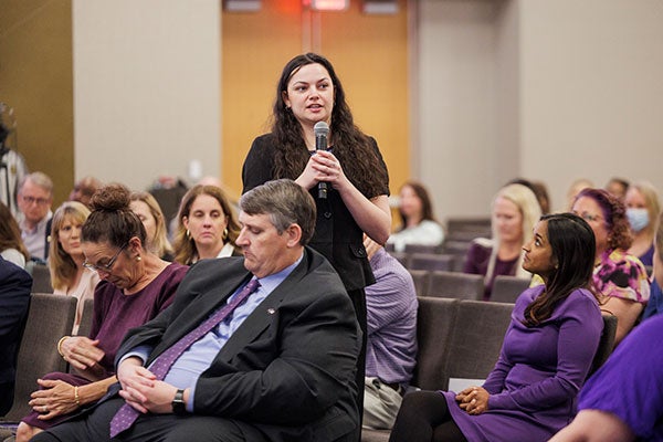 A woman with long black hair stands while talking into a microphone that she is holding while people sitting around her listen.