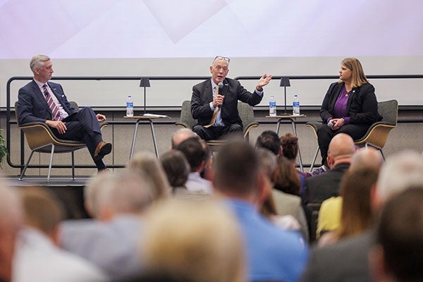 Two men and a woman sit on a stage while the man in the middle gestures with his hand while speaking in a microphone.