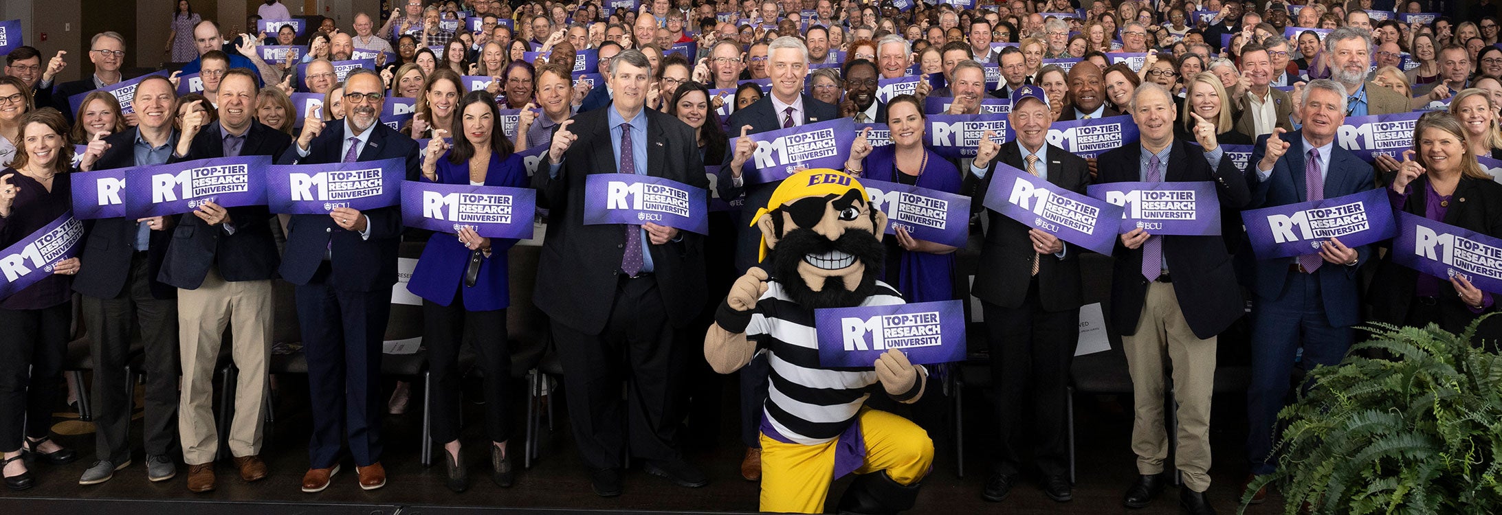 A large group of people, with PeeDee the Pirate kneeling in front, hold up purple Research One signs and make a Pirate hook.