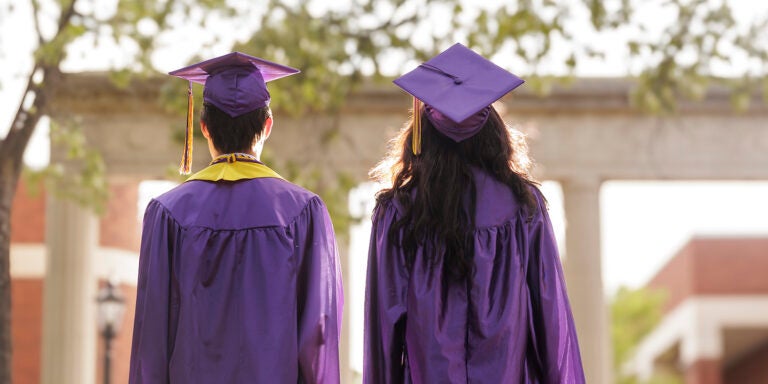 Two people with their back to the camera wear purple graduation caps and gowns.