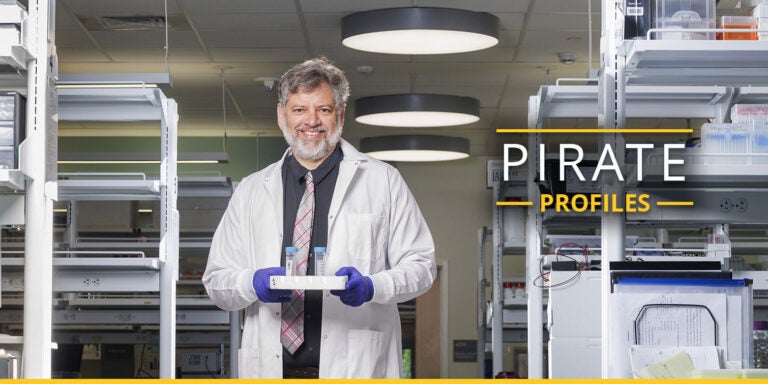 A man wearing a white coat and tie stands in a lab holding a tray of test tubes.