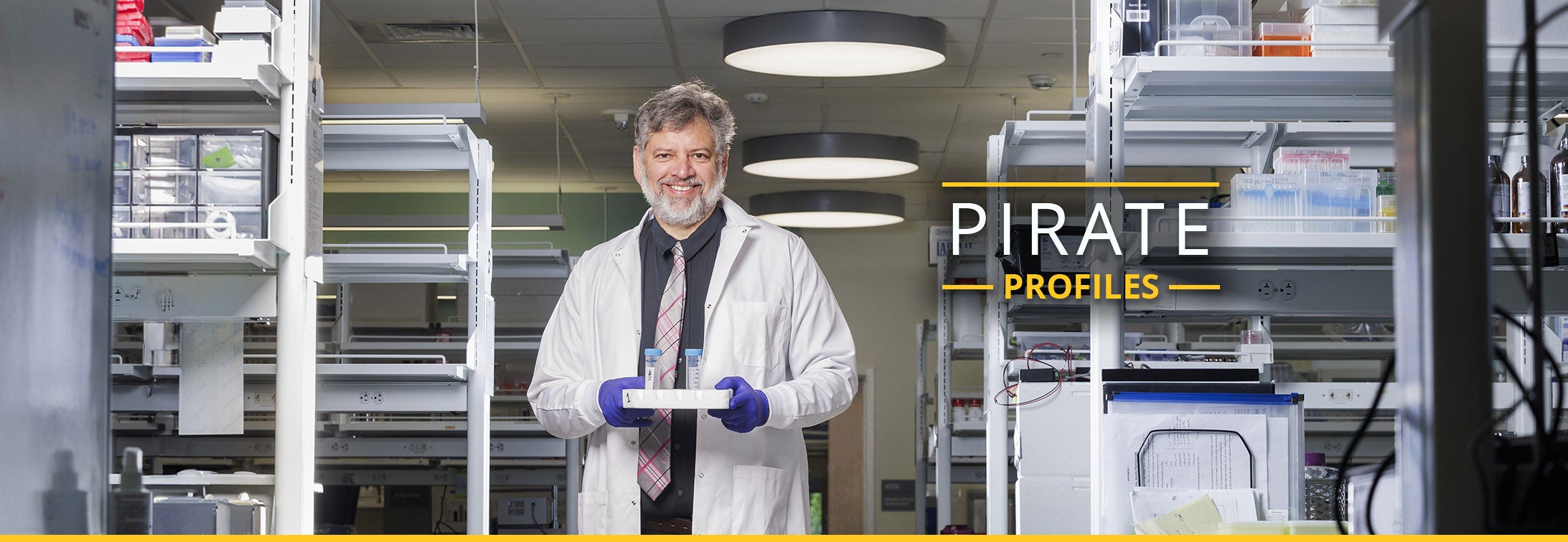 A man wearing a white coat and tie stands in a lab holding a tray of test tubes.