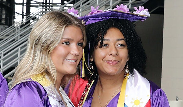 A young woman wearing graduation attire smiles and stands next to another young woman wearing graduation attire.