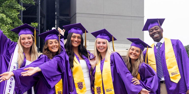 A group of four young women and a young man wearing graduation attire celebrate together.