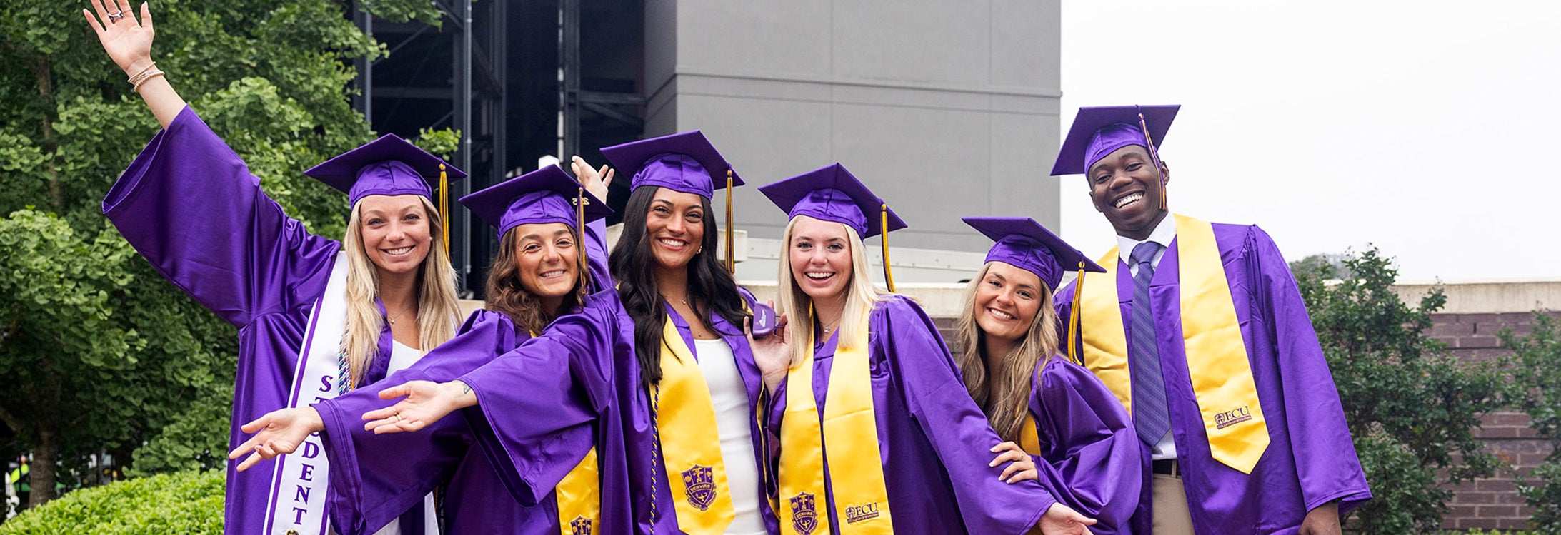 A group of four young women and a young man wearing graduation attire celebrate together.