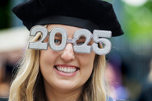 A smiling young woman in graduation attire wears 2025 glasses.