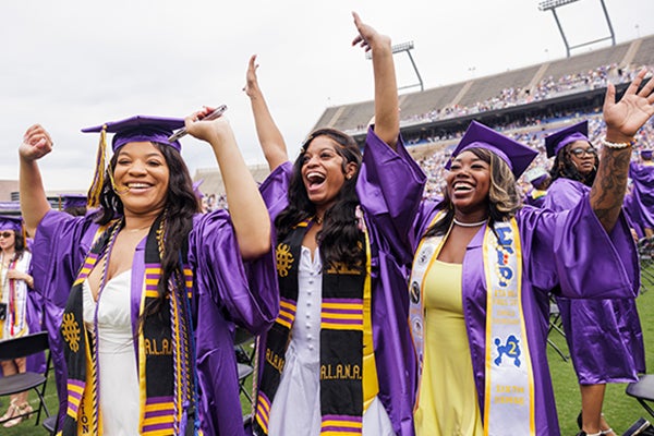 Three young women wearing graduation attire smile with their hands raised in celebration.