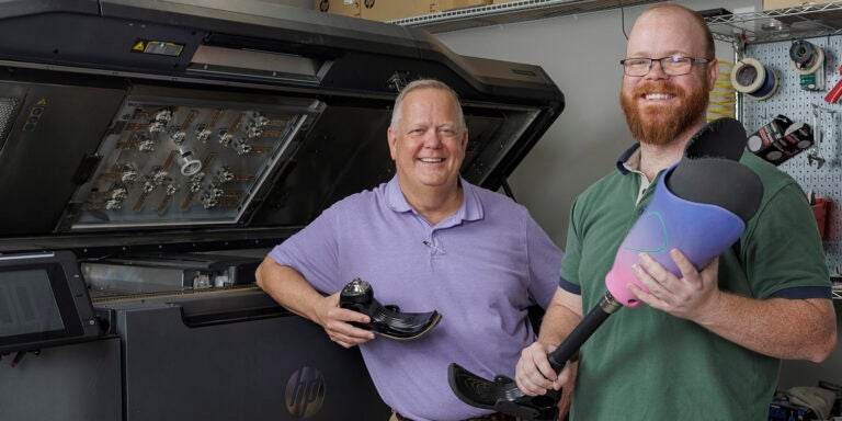 Two men, one at left in a purple shirt and the other in a green shirt, stand next to a large machine that prints three-dimensional objects while holding prosthetic foot attachments.