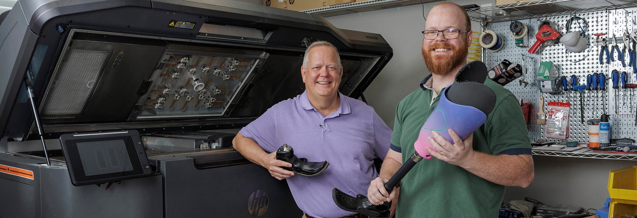 Two men, one at left in a purple shirt and the other in a green shirt, stand next to a large machine that prints three-dimensional objects while holding prosthetic foot attachments.