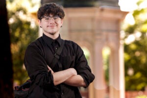 A young man wearing glasses and a black shirt crosses his arms while smiling.