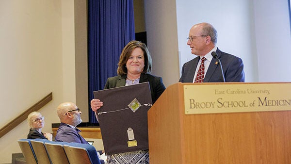 Researcher Dr. Kim Briley stands with N.C. State Representative Dr. Tim Reeder at a podium as Briley’s presented a framed Order of the Long Leaf Pine award.