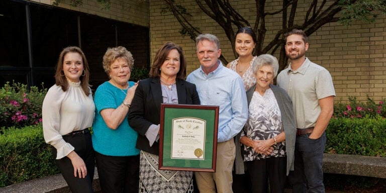 Kim Briley and six of her family members smile and pose with her framed Order of the Long Leaf Pine award outside the Brody Medical Sciences Building.