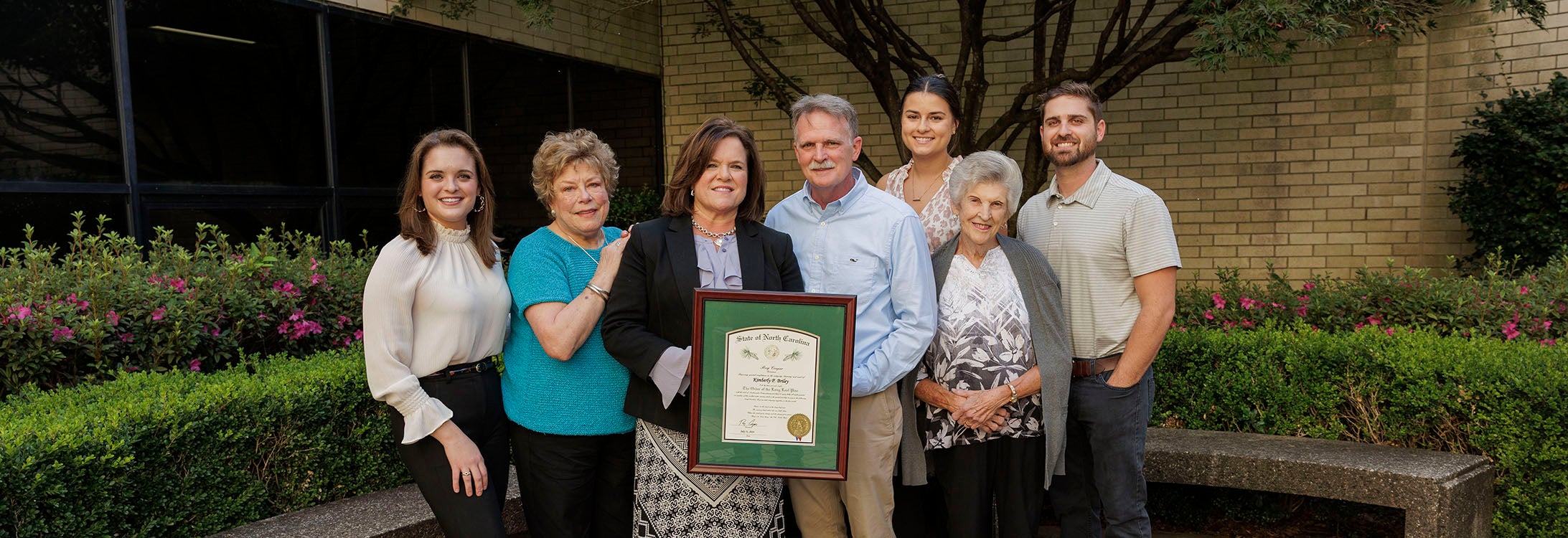 Kim Briley and six of her family members smile and pose with her framed Order of the Long Leaf Pine award outside the Brody Medical Sciences Building.