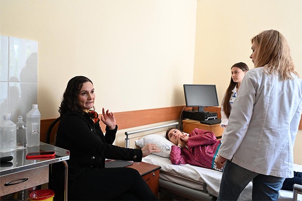 A female nursing instructor in black jacket and pants sits next to a patient in a bed while talking to two female students in white lab coats.