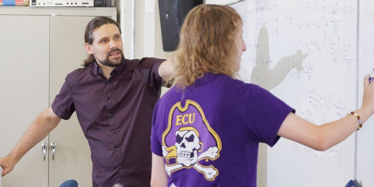 : A man in a maroon shirt points to a white board as another man with his back to the camera writes on the board.