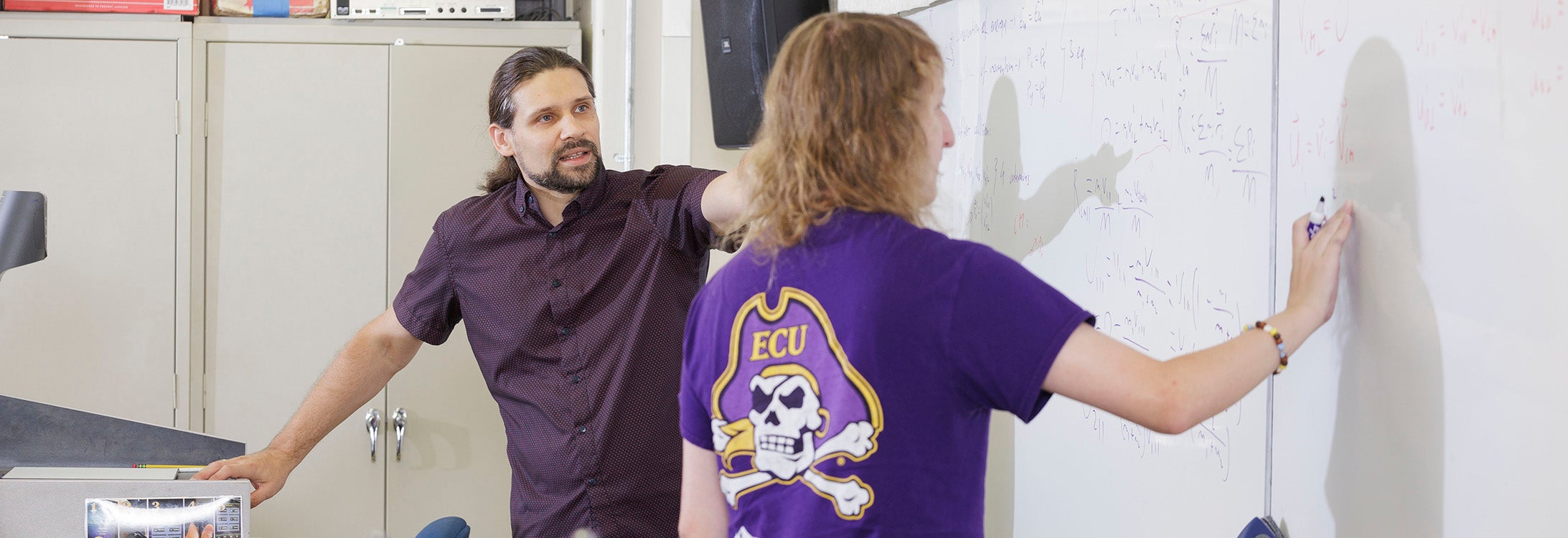 : A man in a maroon shirt points to a white board as another man with his back to the camera writes on the board.