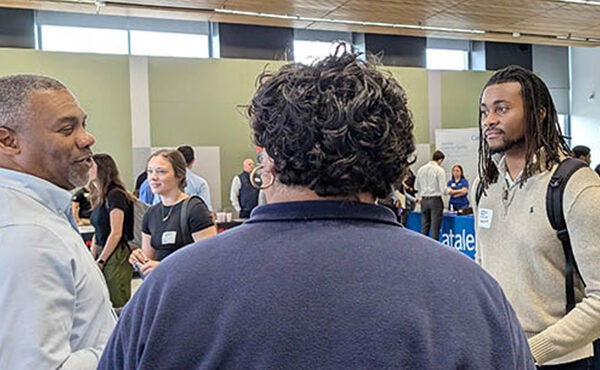 A man in a brown sweater at right stands and talks to two other men, one with his back to the camera and another on the left wearing a blue shirt.