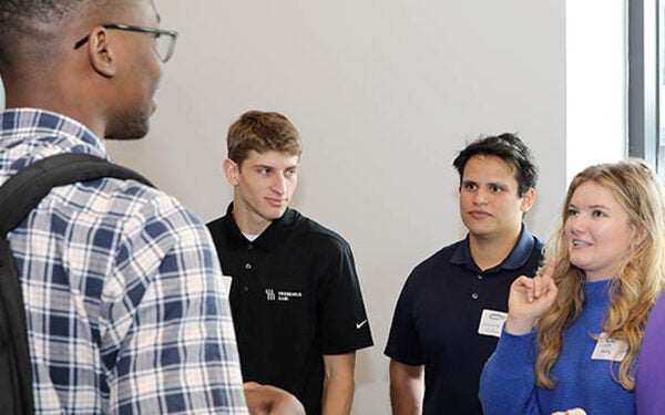 A woman at far right in a blue shirt stands next to two other men in black shirts as they talk to a taller person with his back to the camera.