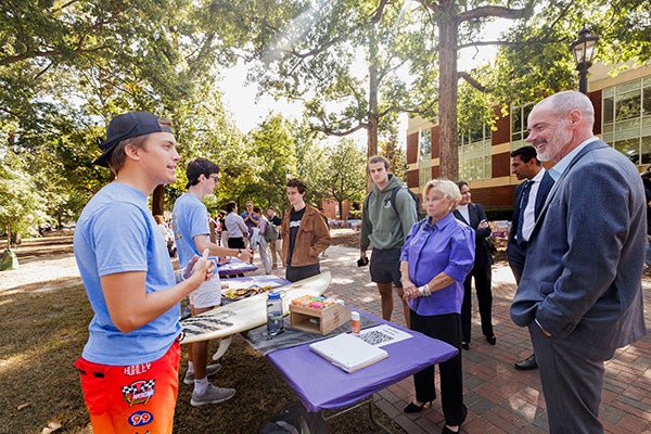 People stand at a table with a surfboard on it while student in a blue shirt and red shorts gestures with his hands.