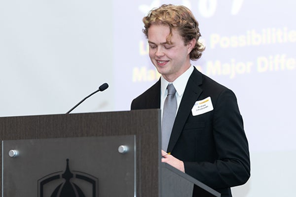 Andrew Jakobowski, wearing a white shirt, gray tie, and black suit jacket, smiles as he looks down and stands behind a podium with the ECU logo and a microphone.