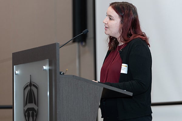 Michaela Trank-Shelp, wearing a maroon top with a black, long-sleeve open-buttoned jacket, stands at a podium with the ECU logo and a microphone.