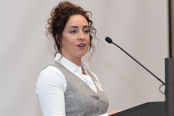 Victoria Hardy-McGowen, wearing a white shirt and gray, striped vest, stands at a podium with a microphone.