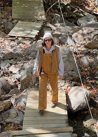 A woman in tan work overalls and a gray sweatshirt walks across a boardwalk in a riverbed during the daylight hours